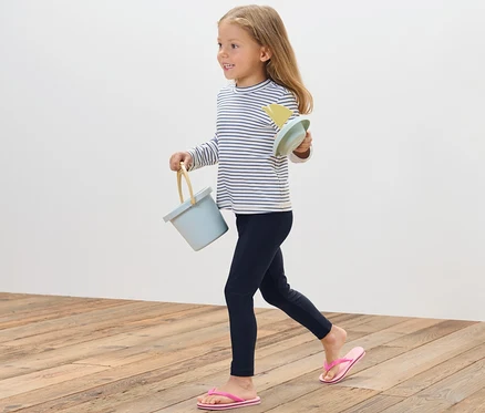 Une fille marche sur un plancher en bois et porte un seau bleu et des jouets de sable. Elle porte un legging de bain pour enfant avec un indice de protection UV 80.