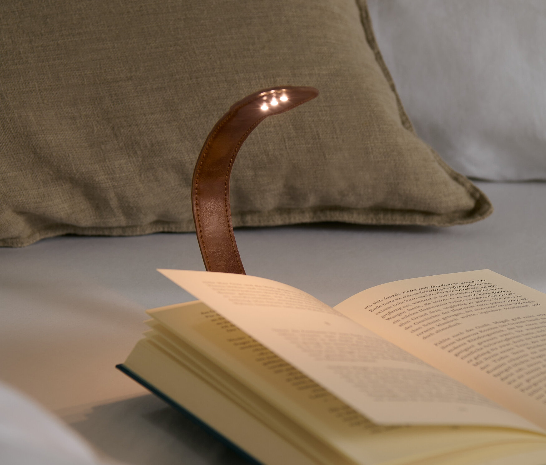 Une lampe de lecture avec clip éclaire un livre ouvert sur un lit avec des coussins.
