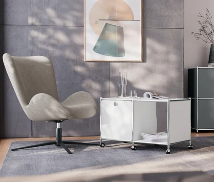 Dans un salon, un fauteuil pivotant est placé à côté d'une table d’appoint métallique « CN3 » blanche avec 1 abattant et roulettes. À l'arrière-plan se trouvent un buffet en métal « CN3 » gris à 2 abattants et une lampe de table en métal chromée.