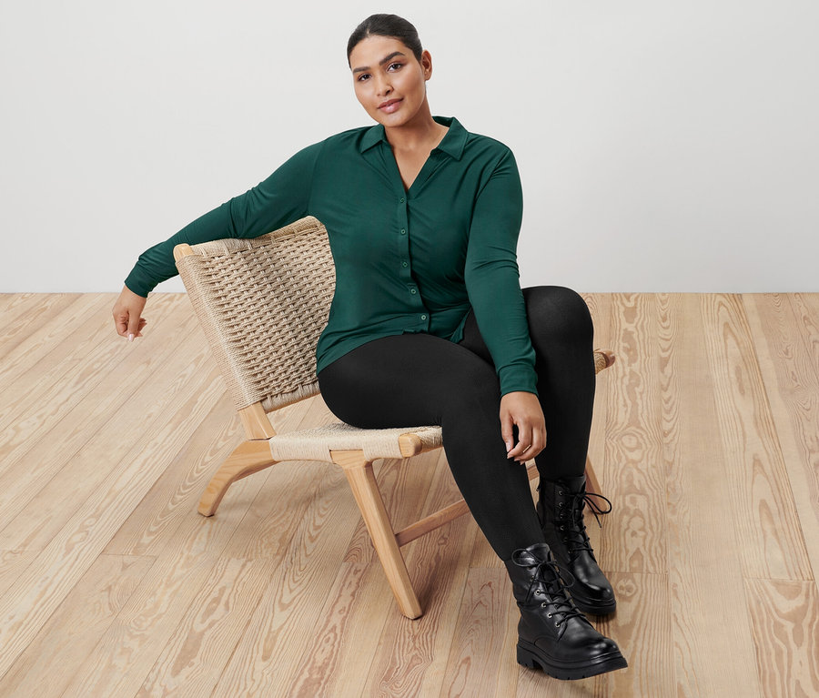 Une femme est assise sur une chaise en osier et porte une blouse verte avec une patte de boutonnage et un legging noir.