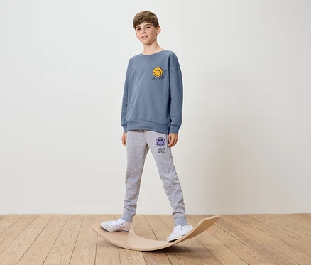 Un garçon portant un sweat-shirt bleu avec un imprimé smiley et un pantalon de survêtement gris se tient sur une planche d'équilibre.