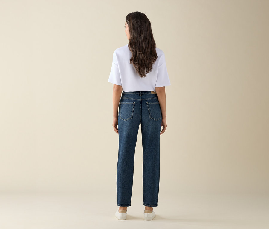 Une femme vue de dos porte un Mom Jeans – Coupe « Nyla », bleu foncé, un t-shirt blanc et des baskets.