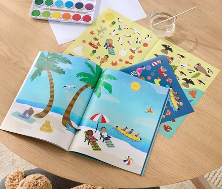 Un Mein Stickerbuch « Au bord de la mer » ouvert avec des illustrations de plage, des couleurs pour peindre et des planches d'autocollants sur une table en bois.