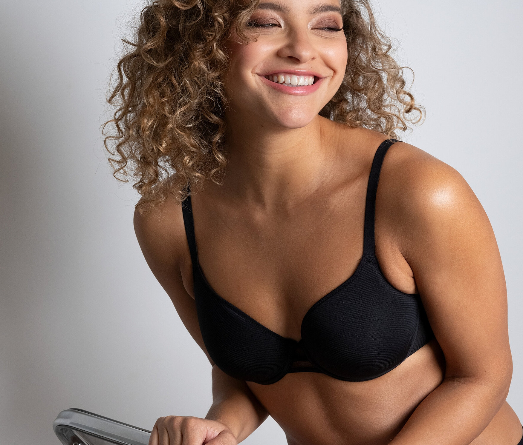 Une femme souriante est assise sur une chaise transparente et porte un soutien-gorge spacer noir « Nais » de Passionata.