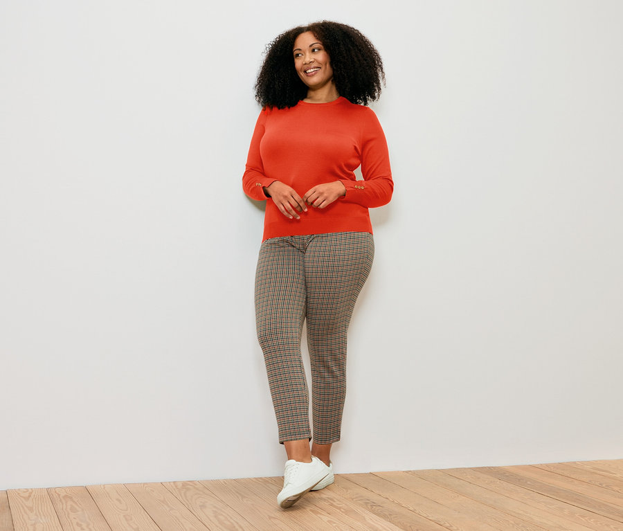 Une femme pose portant un pull en maille fine orange, un pantalon à motifs et des chaussures blanches.