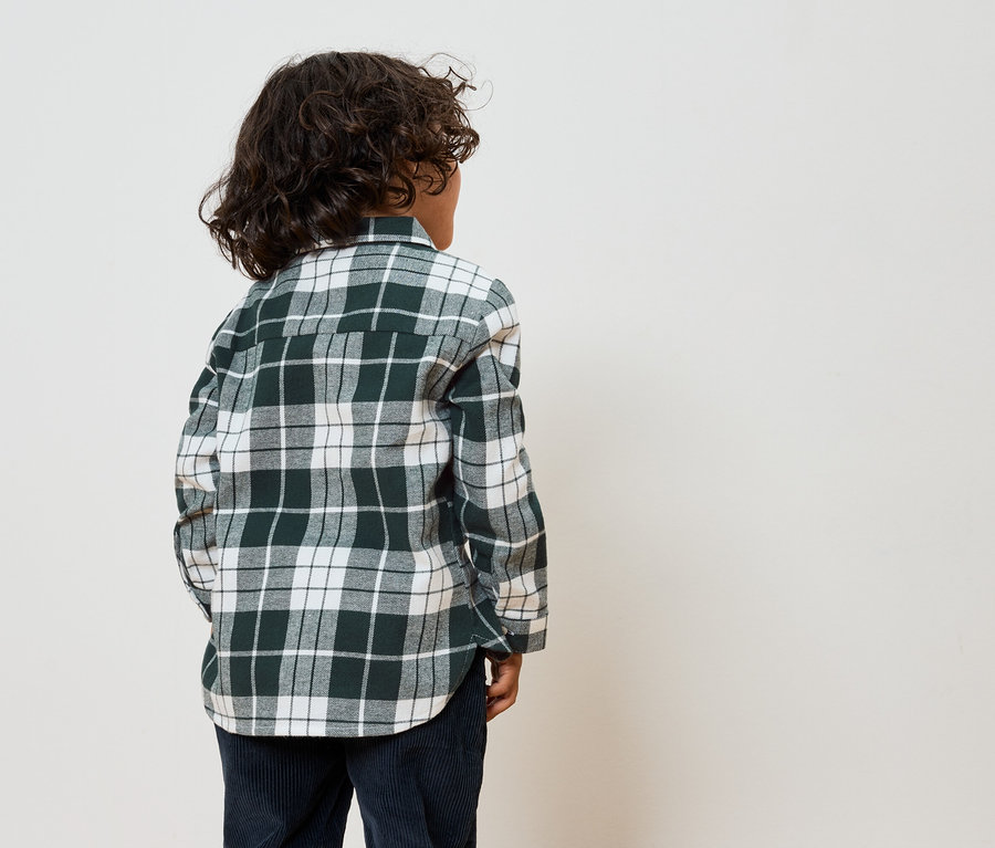 Garçon vu de dos portant une chemise à carreaux verte et blanche et un pantalon bleu marine.