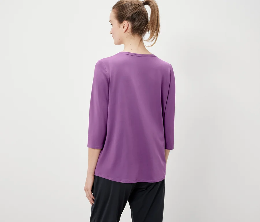 Vue de dos, une femme aux cheveux attachés en queue de cheval porte un t-shirt violet à manches trois-quarts et un pantalon noir.