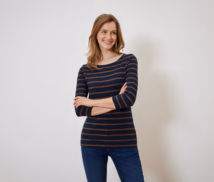 Une femme souriante pose, les bras croisés, portant un t-shirt rayé bleu marine et marron et un jean.