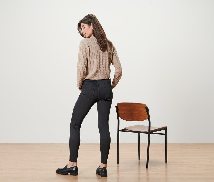Une femme pose en pull beige et jean noir à côté d'une chaise.