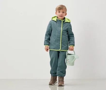 Un garçon en veste et pantalon tient un arrosoir vert clair.
