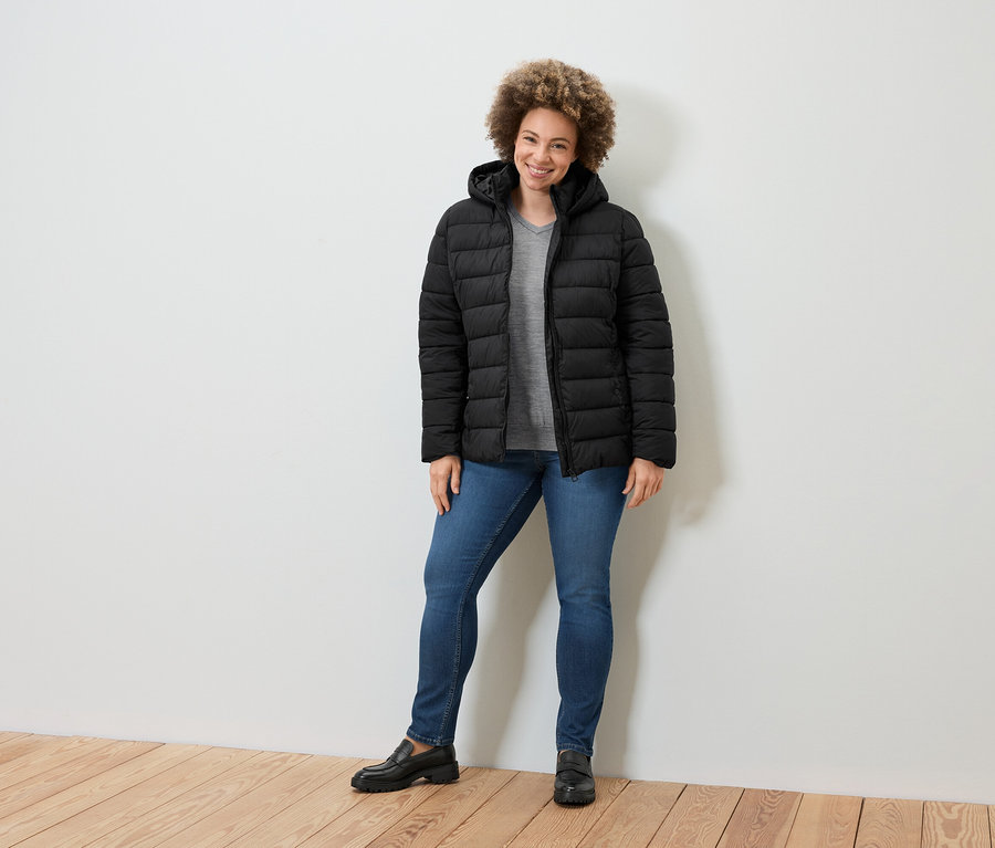 Une femme debout souriant portant une doudoune noire, un jean bleu et des mocassins noirs.