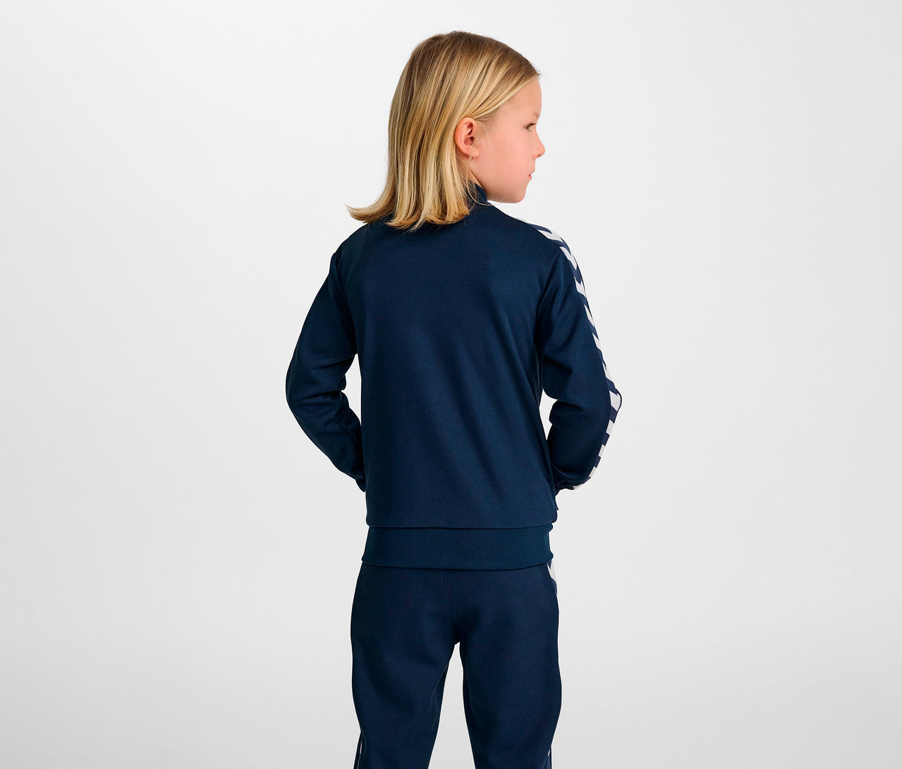 Une fille blonde, vue de dos, porte une veste à fermeture éclair HUMMEL HMLJR Archive Reg Poly bleue.