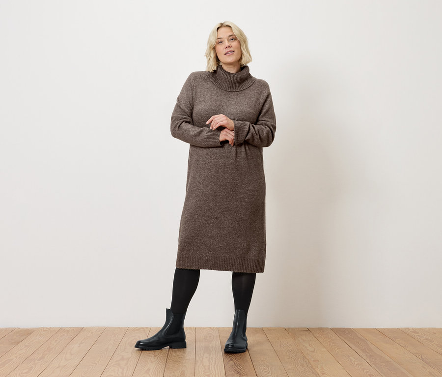 Une femme blonde pose en robe marron à col roulé, legging noir et bottes sur un plancher en bois devant un mur blanc.