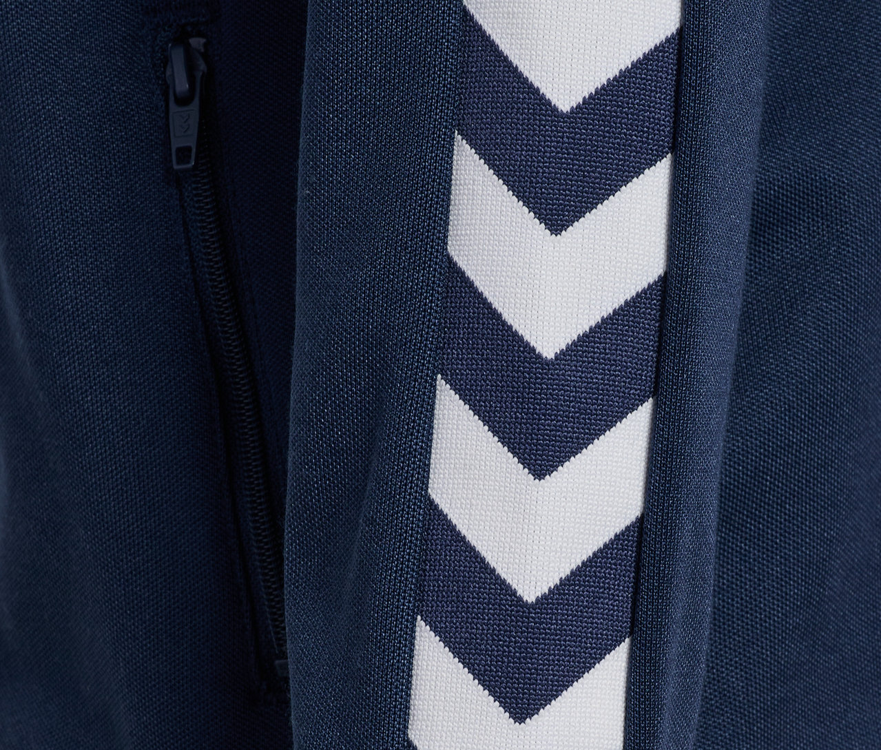 Détail d'une veste zippée bleue Hummel HMLJR Archive Reg Poly avec des chevrons blancs sur la manche et une fermeture éclair.