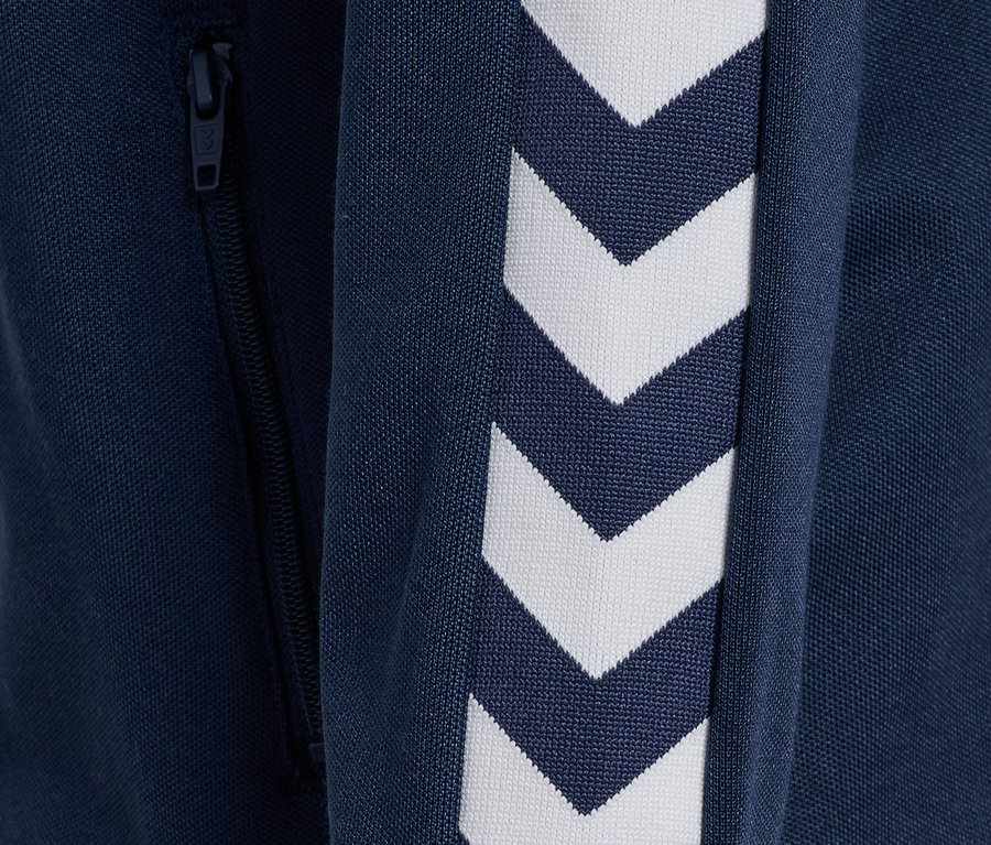 Détail d'une veste zippée bleue Hummel HMLJR Archive Reg Poly avec des chevrons blancs sur la manche et une fermeture éclair.