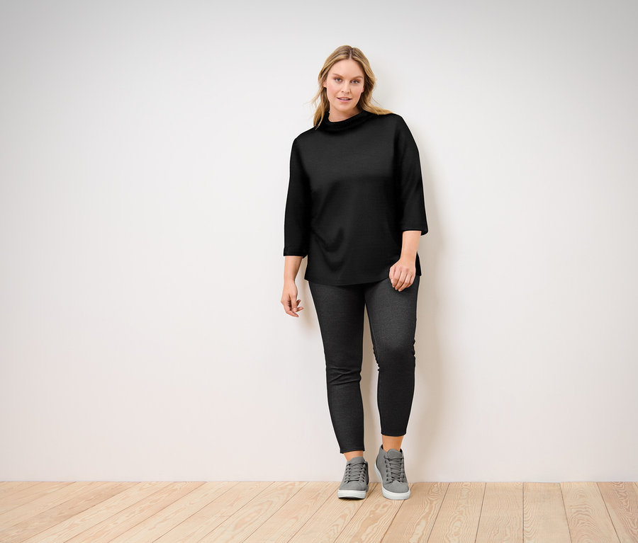 Femme posant dans un haut noir à manches trois-quarts, un jean noir et des baskets grises.