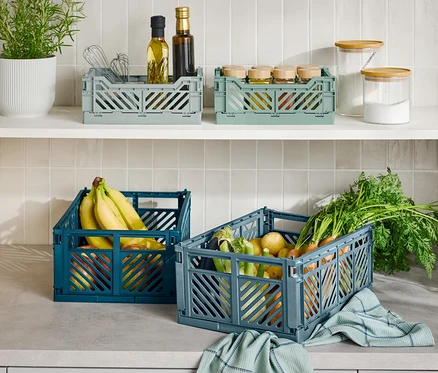 Sur un comptoir de cuisine, trois caisses de rangement pliables, une bleue avec des bananes, une grise avec des légumes et une grise avec des ustensiles de cuisine.