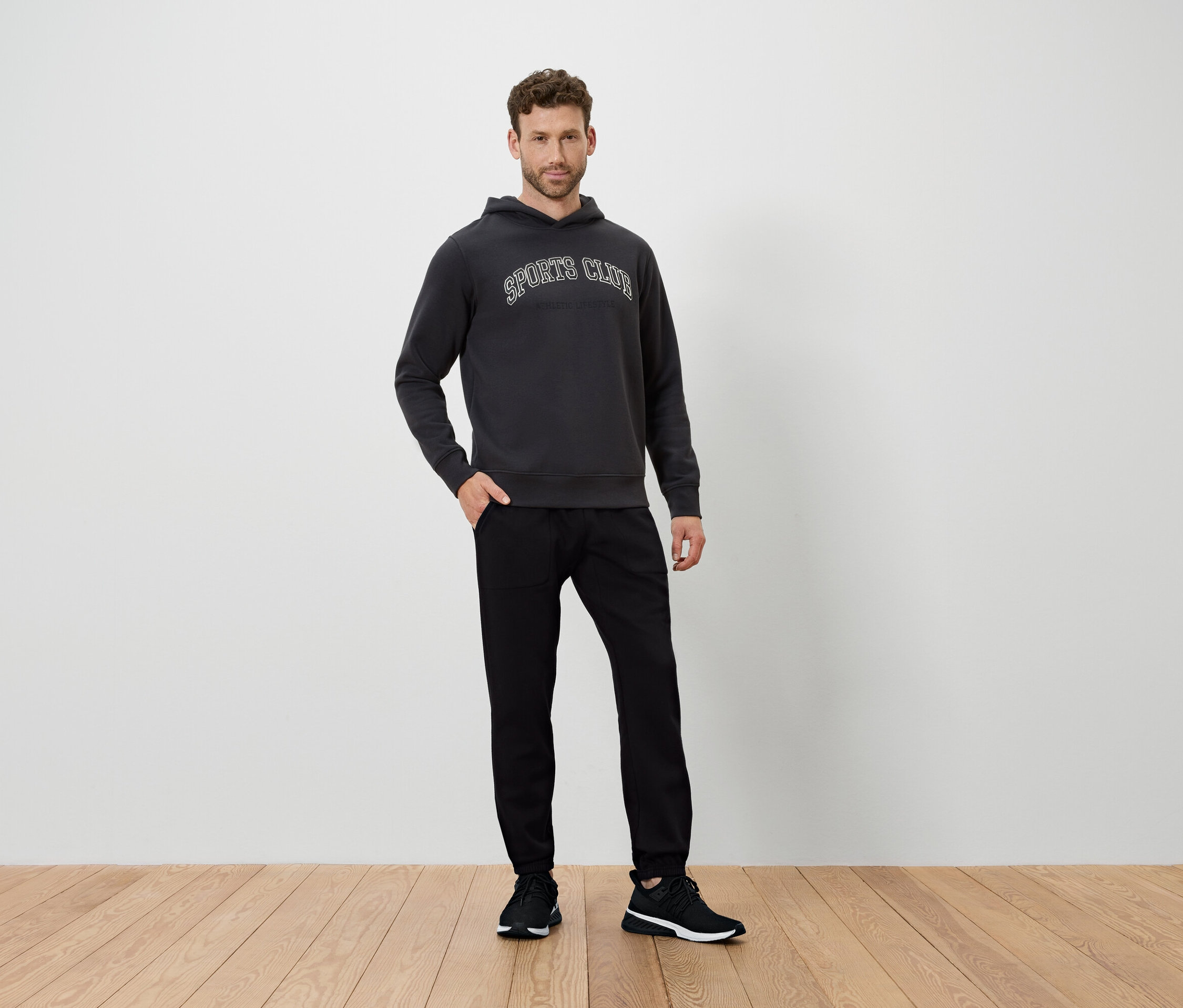 Un homme se tient sur un plancher en bois, vêtu d'un sweat à capuche gris foncé et d'un pantalon noir.