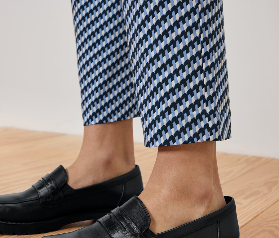 Jambes d'une personne portant un pantalon à motifs bleu et blanc et des mocassins en cuir noirs sur un plancher en bois.