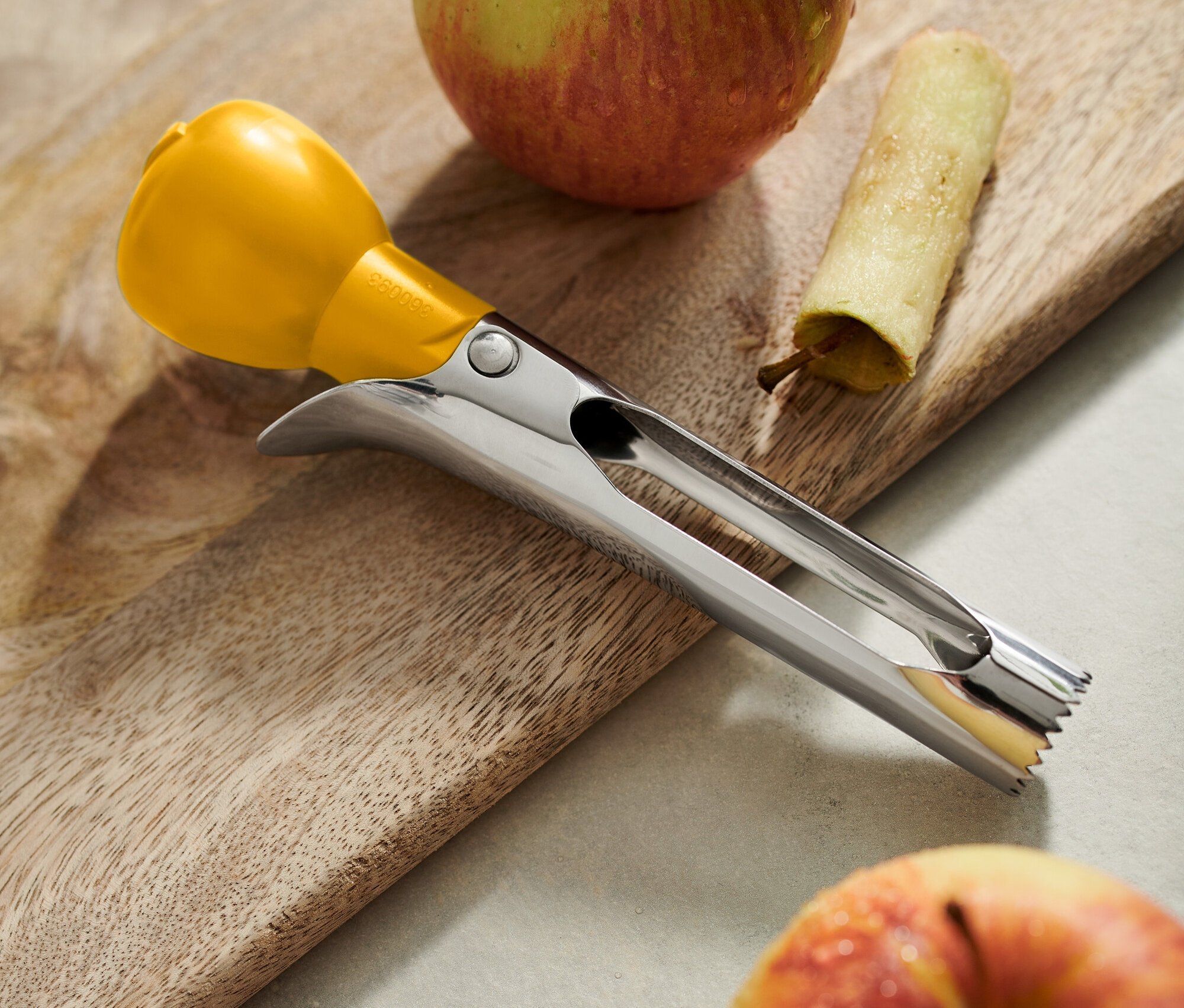 Sur une planche à découper en bois reposent deux pommes, une avec le trognon enlevé, à côté un morceau de peau et un vide-pomme en acier inoxydable avec un manche jaune.