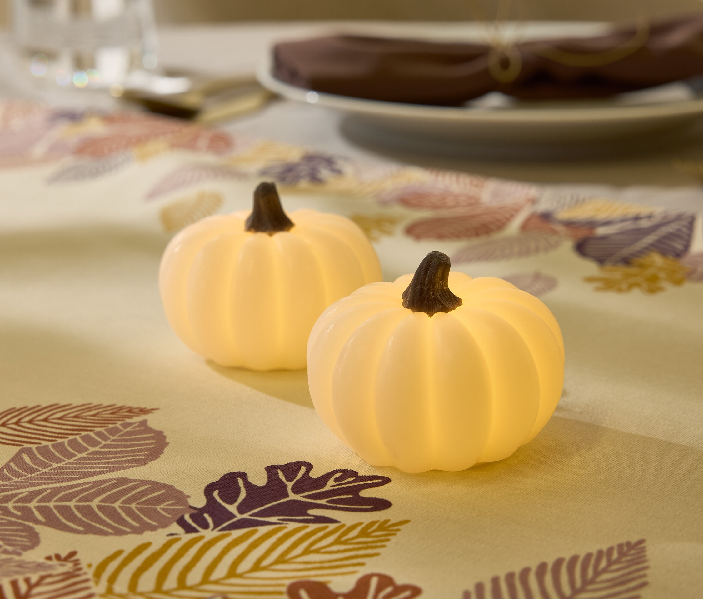 Deux citrouilles blanches illuminent un chemin de table à motifs de feuilles, avec une assiette et un verre en arrière-plan.