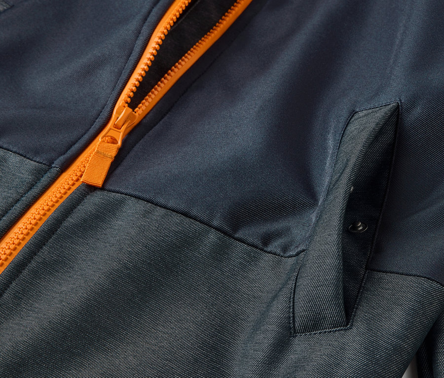 Détail d'une veste en softshell pour enfant avec fermeture éclair orange et poche.