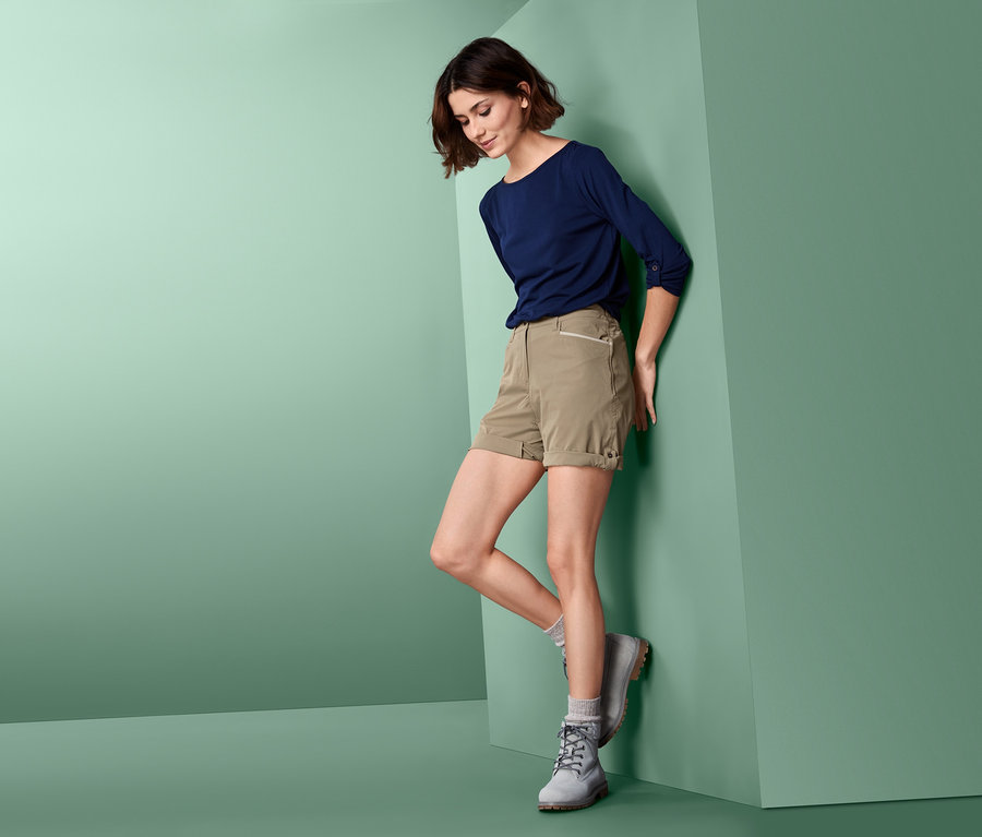 Une femme appuyée contre un mur porte un short fonctionnel en softshell, un t-shirt bleu et des bottes grises.