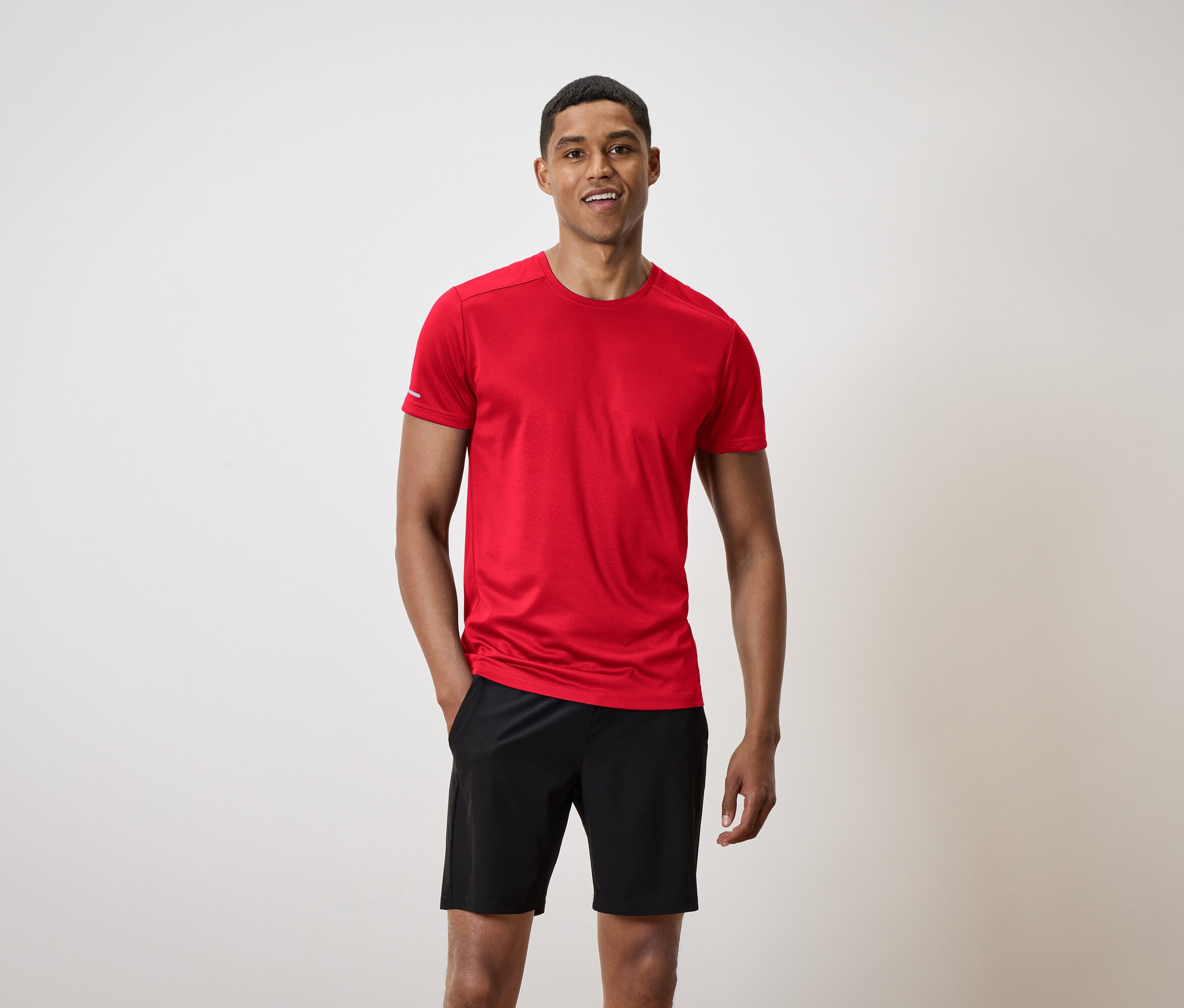Un homme souriant aux cheveux courts se tient debout. Il porte un t-shirt rouge et un short noir.