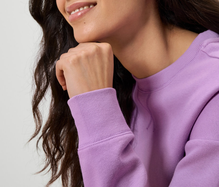 Gros plan sur une femme aux cheveux ondulés et foncés, qui pose sa main sur son menton. Elle porte un pull violet clair.