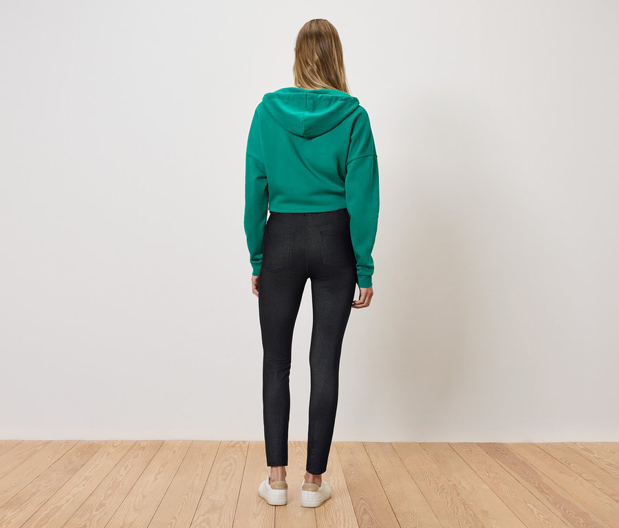 Une femme se tient dos à la caméra, portant un sweat à capuche vert et un legging noir.