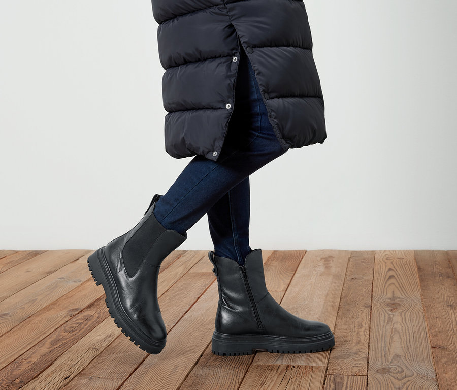 Jambes d'une personne portant un jean et des bottes en cuir noires sur un plancher en bois. La personne porte une longue veste noire.