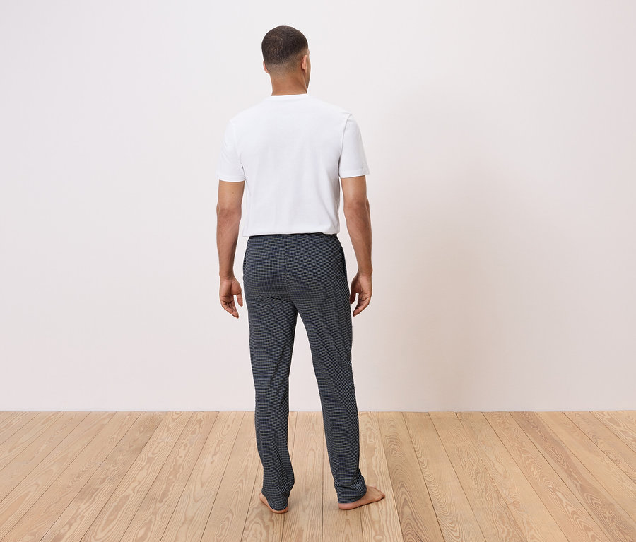 Un homme, dos à la caméra, se tient sur un plancher en bois, portant un t-shirt blanc et un pantalon sombre à carreaux.