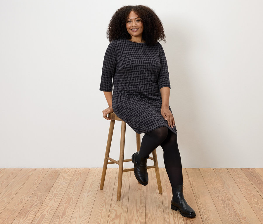 Une femme est assise sur un tabouret et porte une robe en jersey jacquard pied-de-poule.