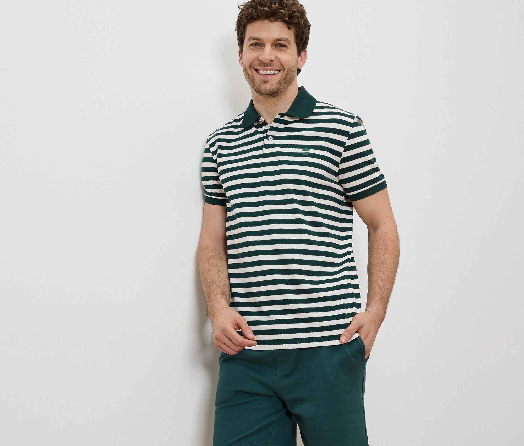 Un homme debout souriant, portant un polo en tissu piqué à rayures horizontales et un short.