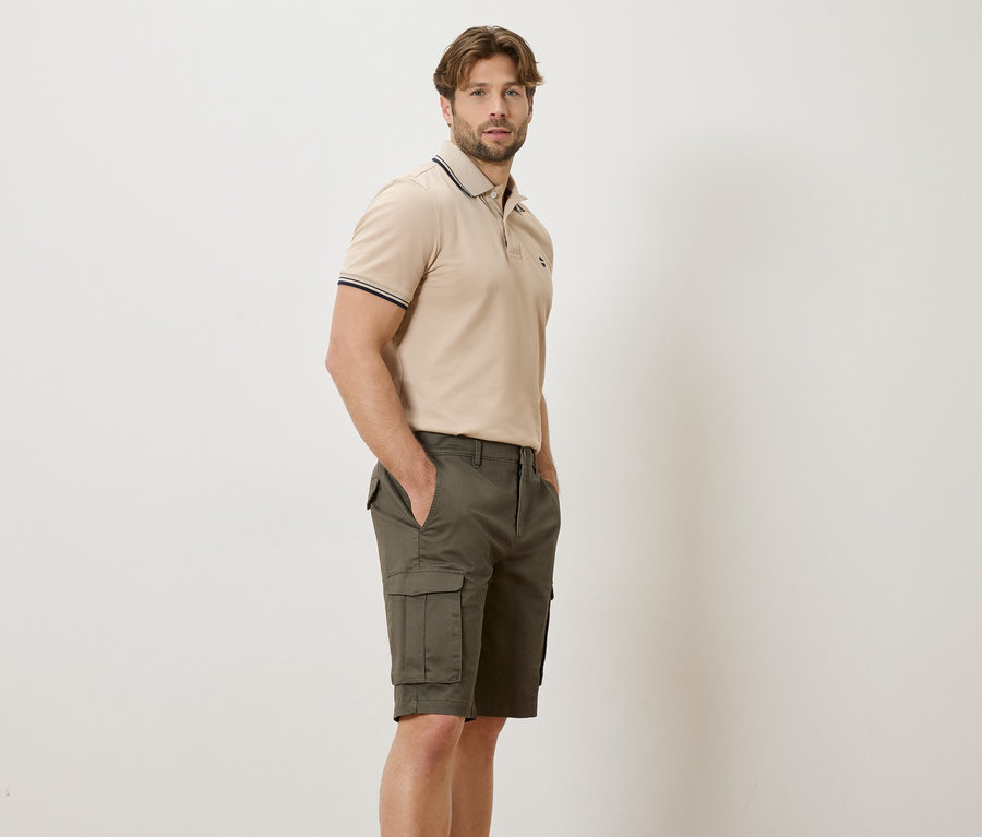 Un homme se tient de profil, portant un polo beige et un short cargo vert sur un fond clair.