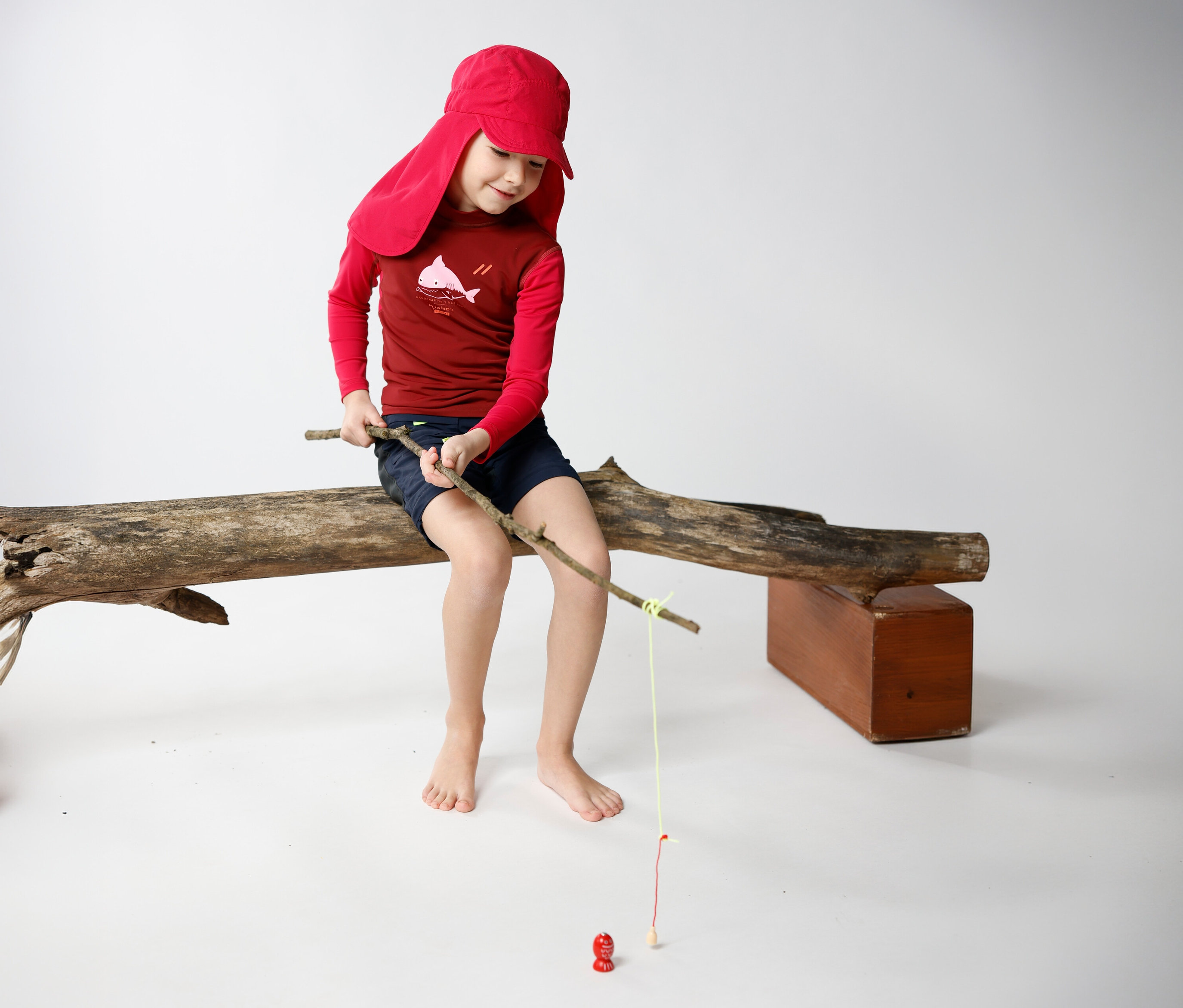 Un garçon assis sur un tronc d'arbre, coiffé d'une casquette JAKO-O avec protège-nuque couleur raisin, pêche avec un bâton.