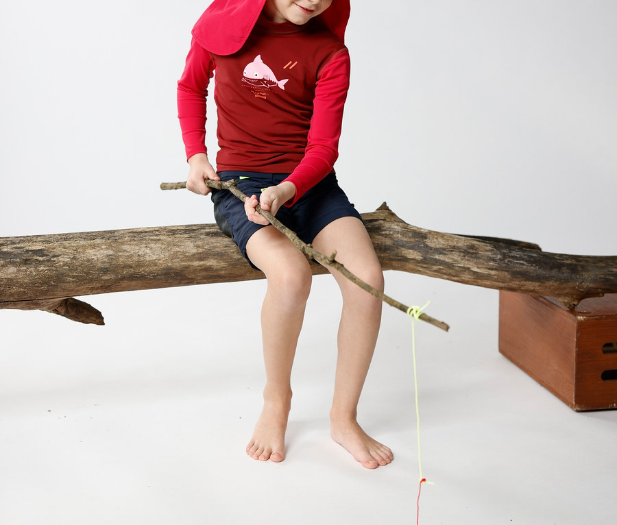 Un enfant avec un chapeau rouge est assis sur un tronc d'arbre et pêche avec un bâton.