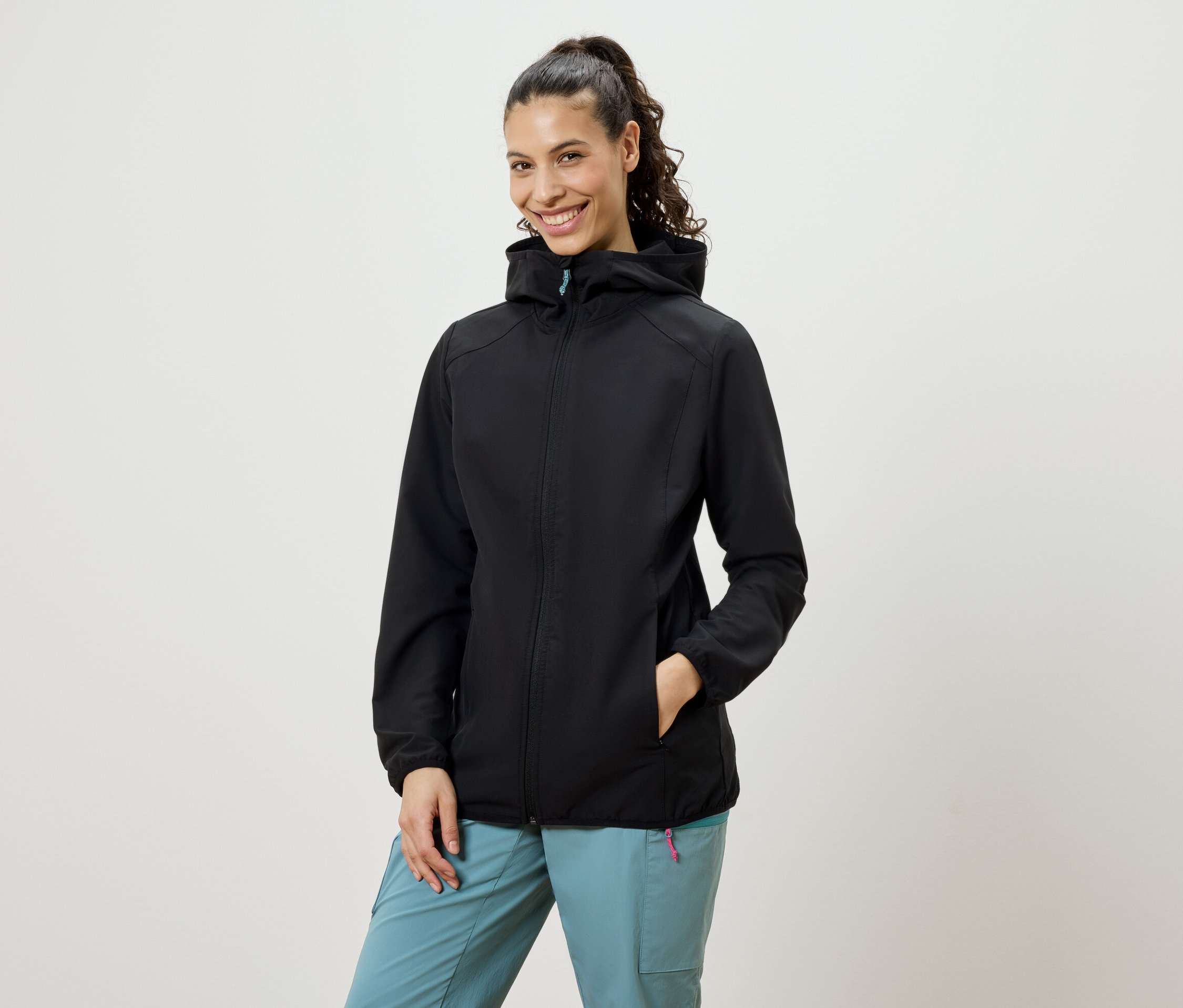 Une femme souriante avec une queue de cheval porte une veste softshell noire et un pantalon bleu.