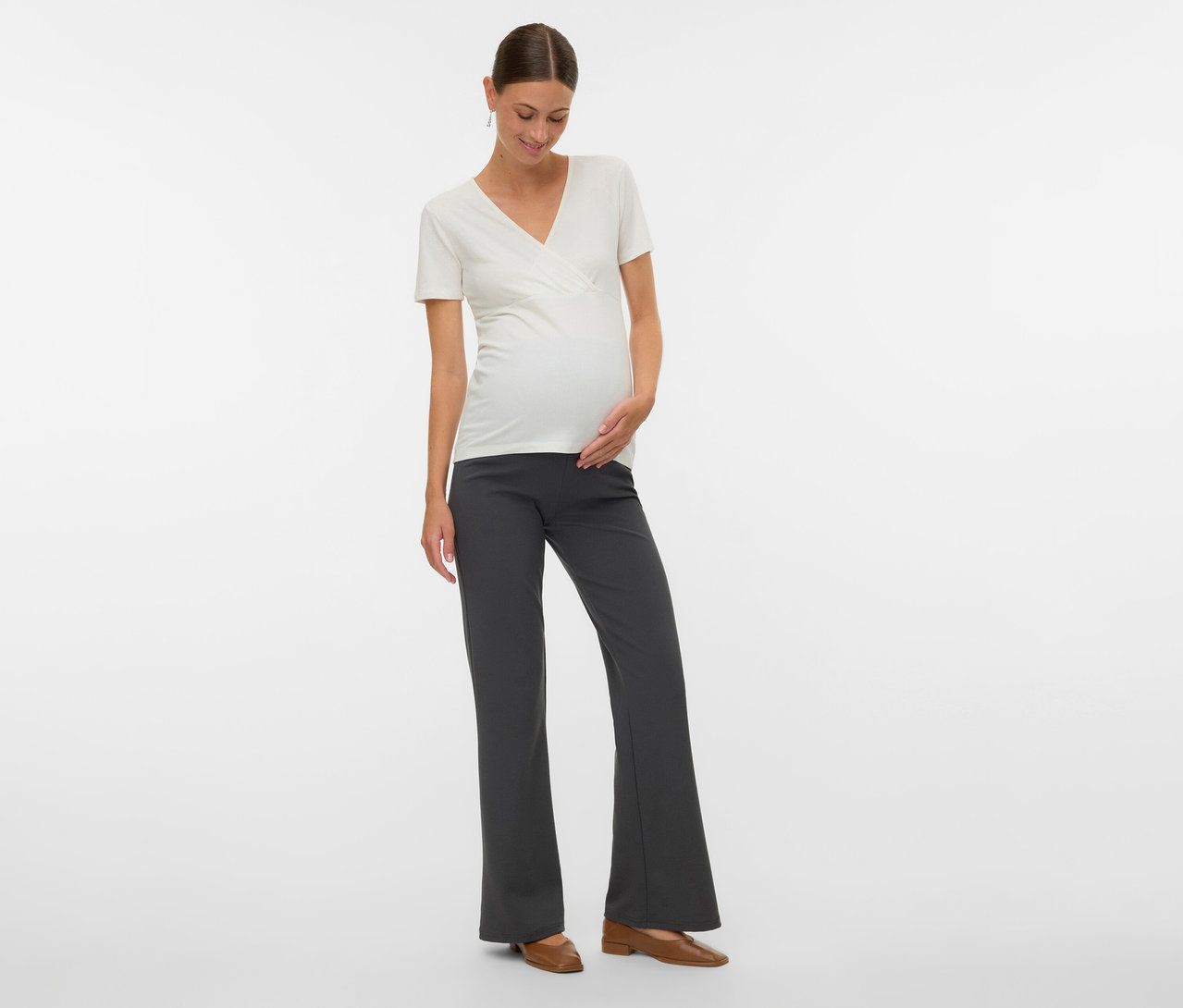 Une femme enceinte porte un lot de 2 t-shirts MAMALICIOUS «
MLKERRIE TESS », un pantalon gris et des chaussures marron.