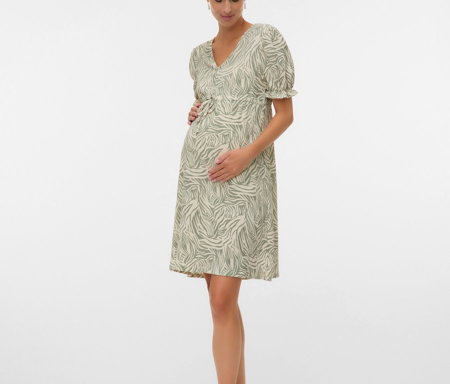 Femme enceinte portant une robe vert et blanc avec des sandales, les mains sur le ventre.