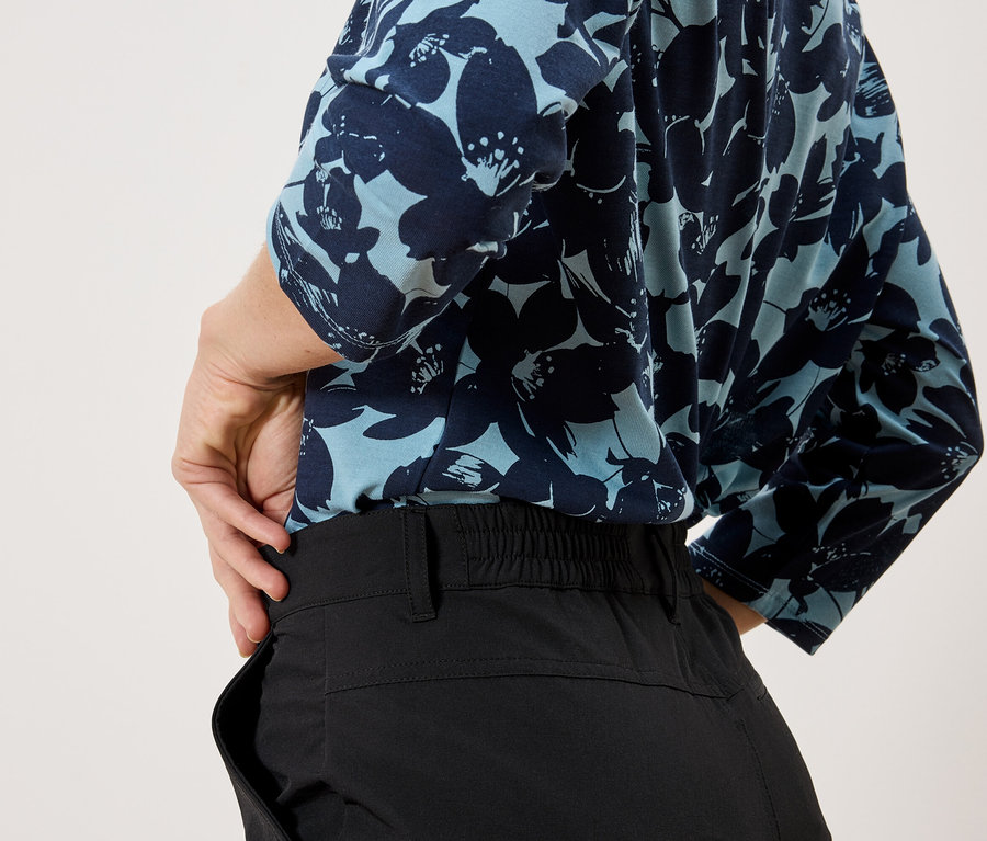Une personne portant un haut bleu clair à motifs floraux et un pantalon noir se tient devant un fond blanc.