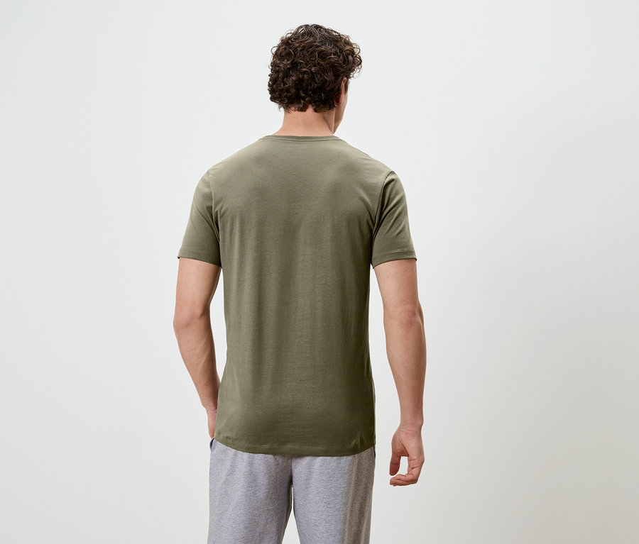 Un homme aux cheveux bouclés se tient dos à la caméra, portant un t-shirt vert olive et un short gris.