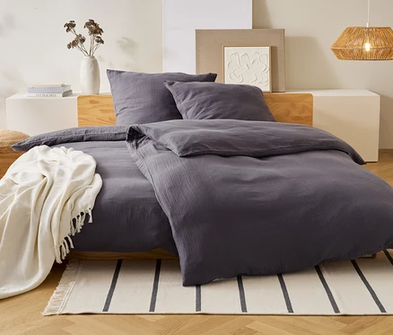 Du linge de lit gris est posé sur un lit avec des oreillers. Une couverture blanche est posée sur le lit. Un vase et une lampe sont à l'arrière-plan.