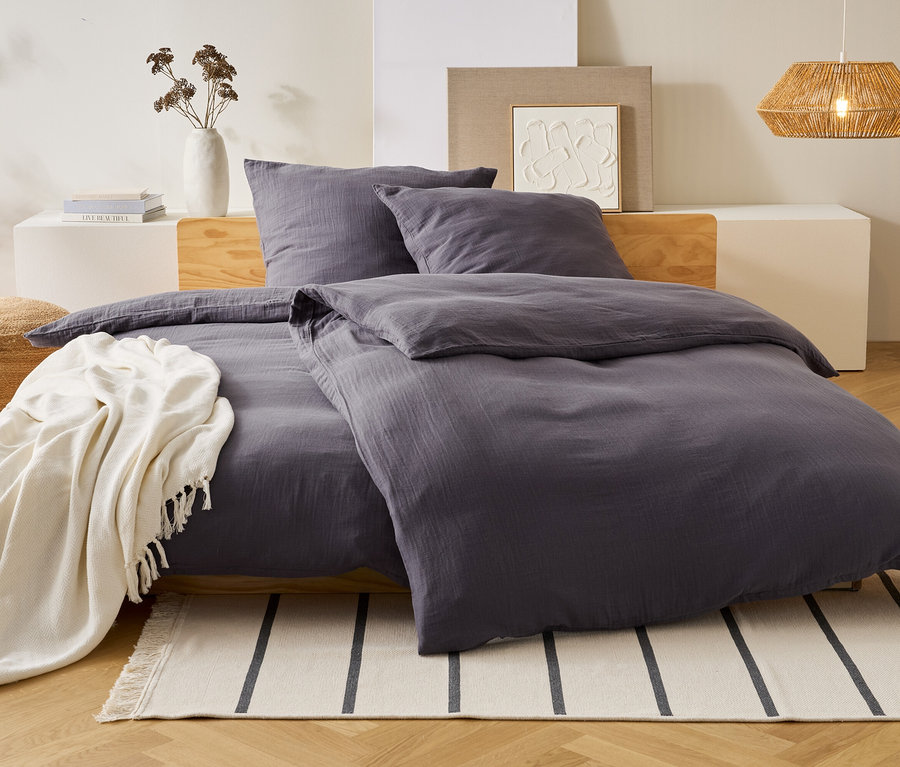 Du linge de lit gris est posé sur un lit avec des oreillers. Une couverture blanche est posée sur le lit. Un vase et une lampe sont à l'arrière-plan.