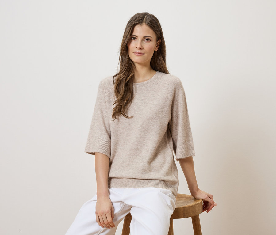 Une femme aux longs cheveux bruns est assise sur une chaise en bois, portant un pull beige et un pantalon blanc.
