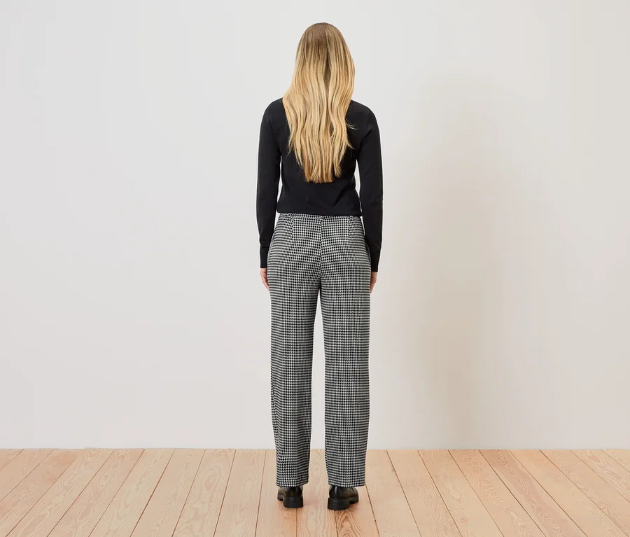 Une femme blonde se tient dos à la caméra, portant un haut noir et un pantalon à carreaux noirs et blancs.
