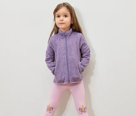 Fillette aux cheveux bruns portant une veste en polaire violette et un legging rose avec un motif de licorne.