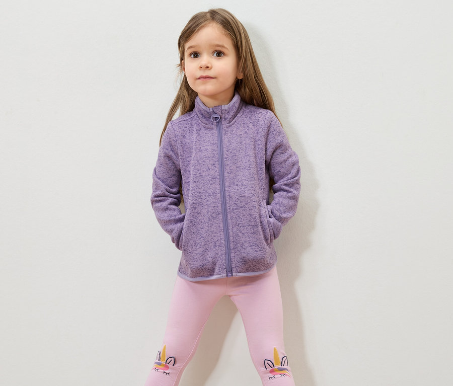 Fillette aux cheveux bruns portant une veste en polaire violette et un legging rose avec un motif de licorne.