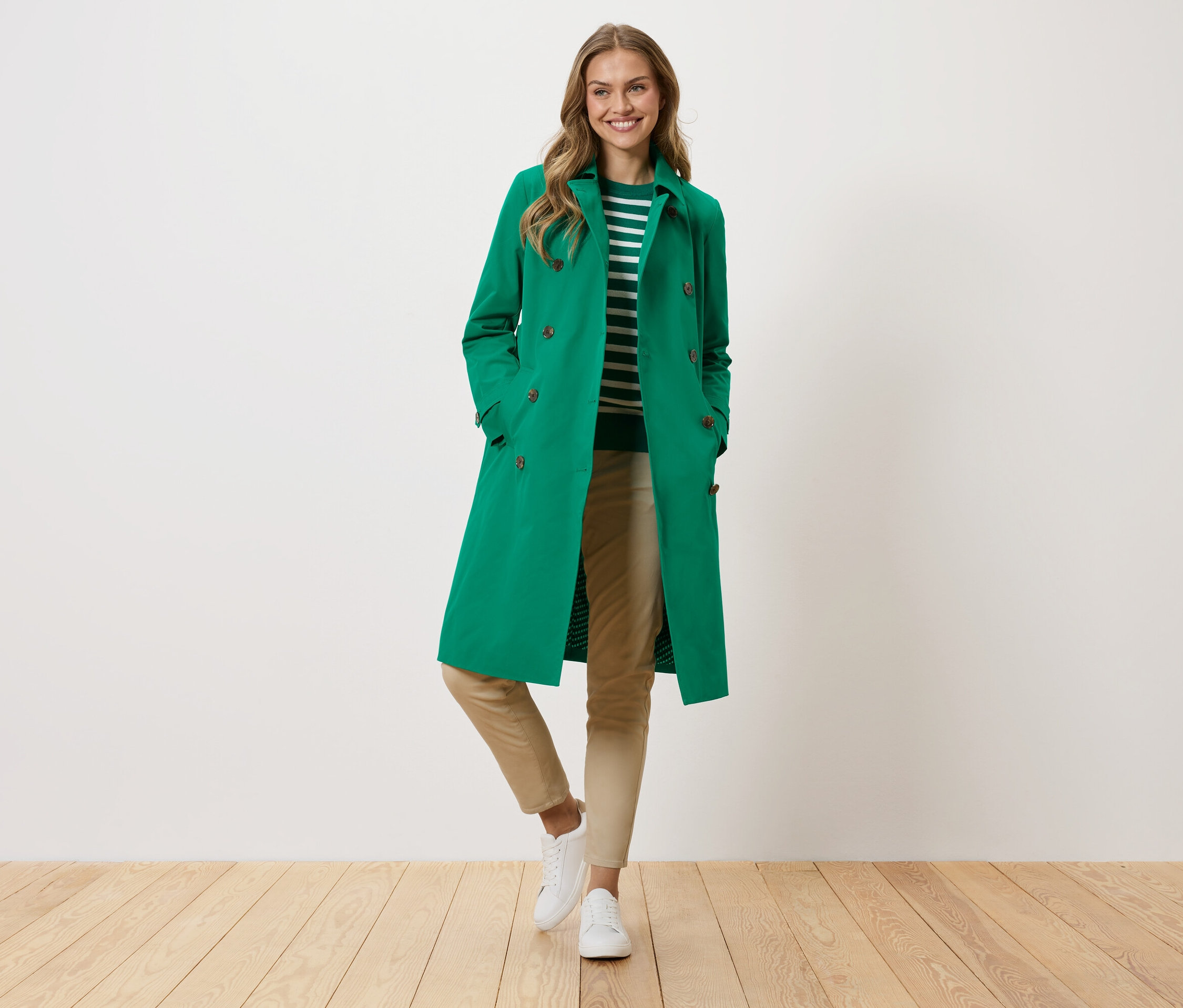 Une femme portant un manteau vert, un pull rayé, un pantalon beige et des baskets blanches se tient devant un mur blanc.