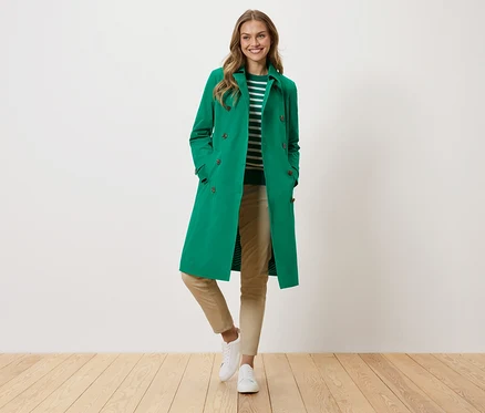 Une femme portant un manteau vert, un pull rayé, un pantalon beige et des baskets blanches se tient devant un mur blanc.